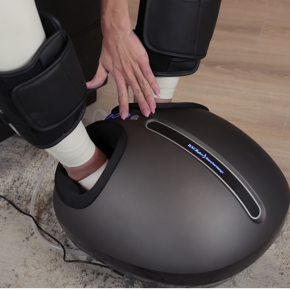 A person adjusts settings on a sleek, shiatsu foot massager while seated. Their feet are inside the device, conveying relaxation and comfort.