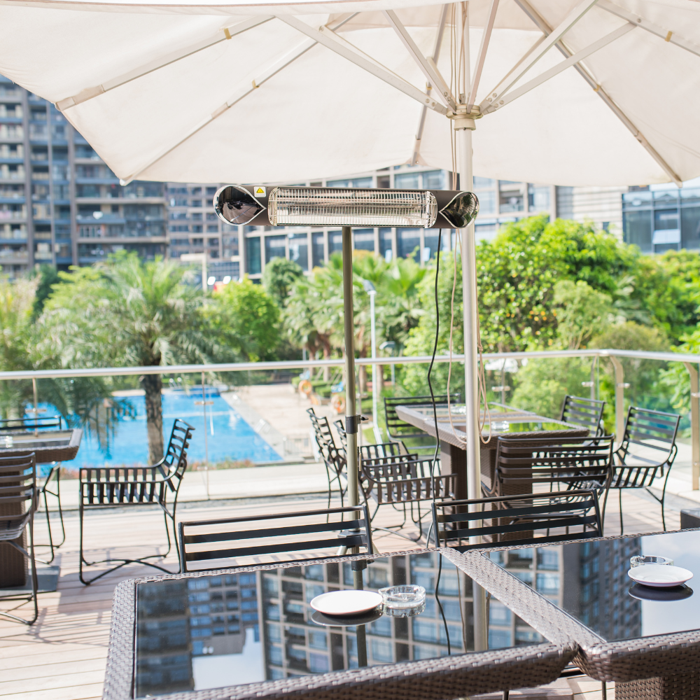 Sunny outdoor cafe with wicker tables topped with glass and metal chairs under large umbrellas. A pool and lush greenery create a relaxing ambiance with an infrared heater.