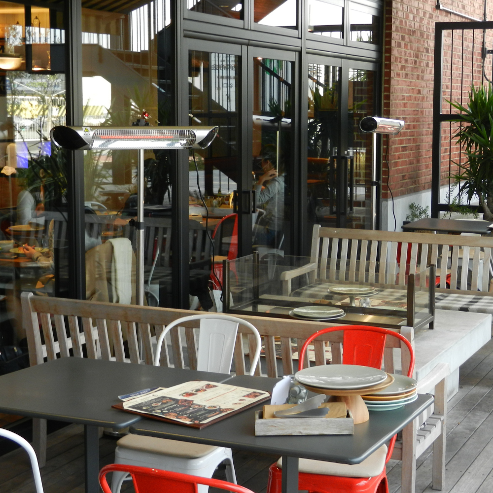 Infrared heaters are providing warmth to an outdoor café scene with modern seating, red and white chairs, metal tables, menus, and stacked plates. Glass walls reveal diners inside.