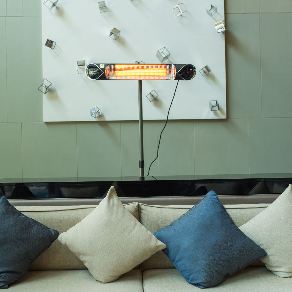 A freestanding iQ Heat® Wave infrared heater is positioned behind a beige sofa with assorted pillows; modern decor with geometric wall art is visible in the background.